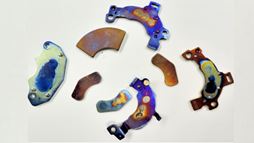 A photo of four voice coil magnets taken from a desktop or laptop computer and their brackets (which have mounting holes).  These magnets are each about 10.5 &ndash; 15 grams, 1 - 1.25 inches long.  These ones are colorful, because they have been overheated in the process of separation and demagnetization. Photo supplied by the Ames Laboratory.