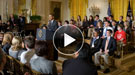 President Obama speaking at a podium with awardees seated behind him and an audience in front of him